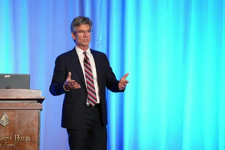 Alexander Green Oxford Communiqué keynote speaker photo – gray-haired man in suit and striped tie presenting on stage at Hiltons Hotel Texas conference, arms outstretched explaining investment strategy, blue curtain background, podium with laptop, live financial education event, bestselling author Alexander Green speaking engagement, stock market trading seminar, high-net-worth investing presentation, Oxford Club live event imagery.