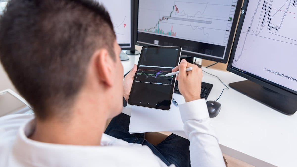 Trader reviewing cryptocurrency charts on tablet with multiple market data screens in background