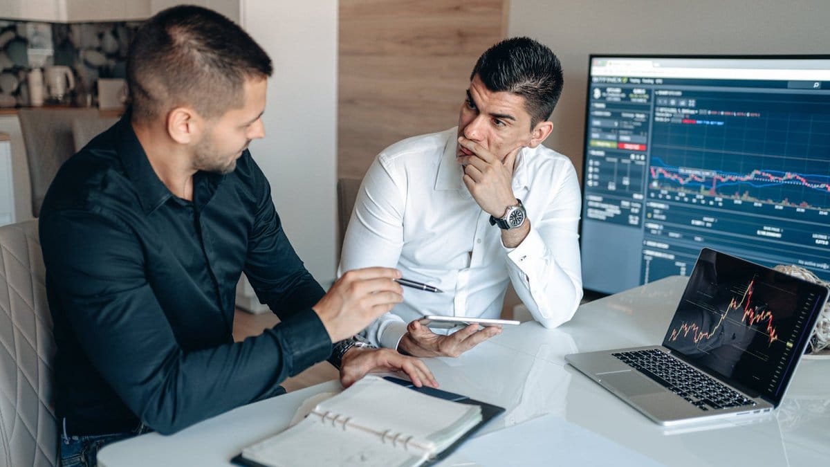 Two financial professionals discussing market strategy with trading charts on screen