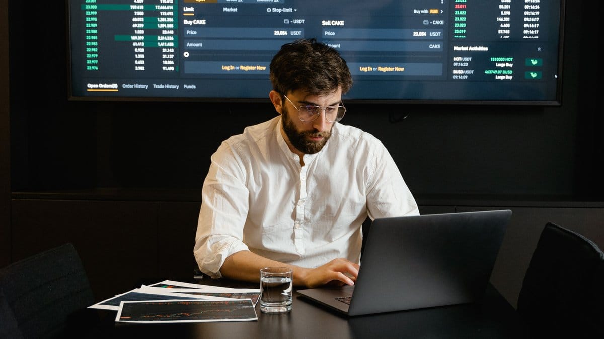 A focused financial professional analyzes stock market trends on a laptop with market data displayed on a large screen behind him.