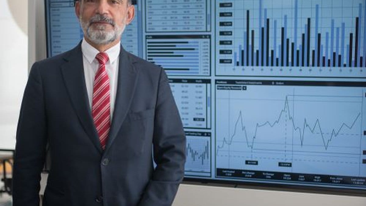 Senior financial executive standing before economic data dashboard showing market indicators