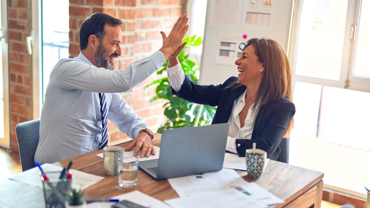 Two professionals celebrating successful retirement planning at a desk with financial documents and a laptop